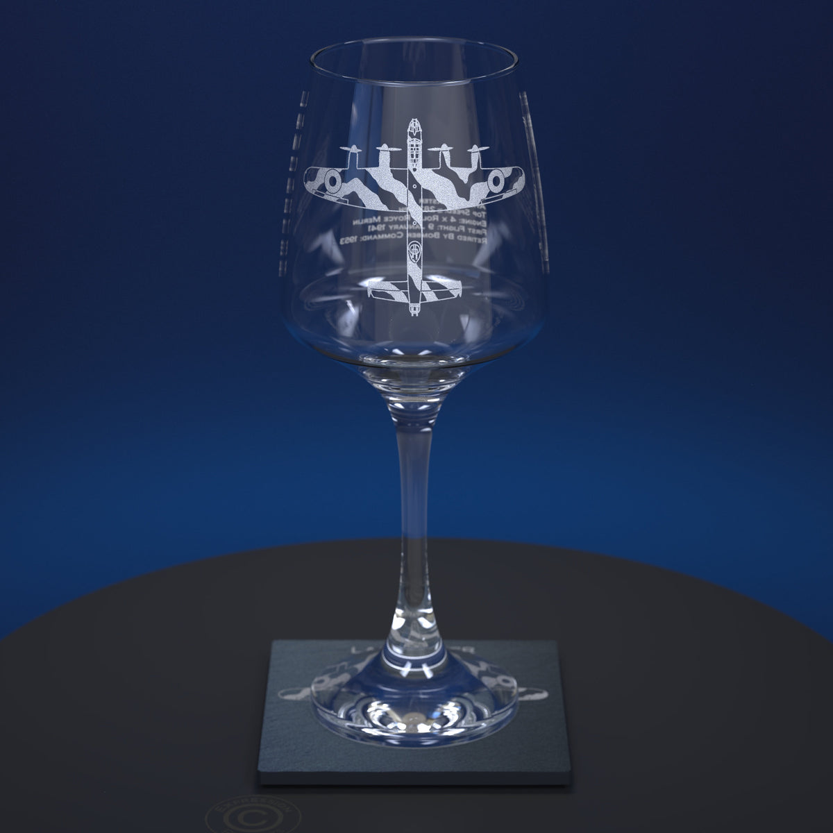 A clear stemmed wine glass with an etched Lancaster aircraft design and text on the bowl, displayed on a dark coaster against a blue gradient background.