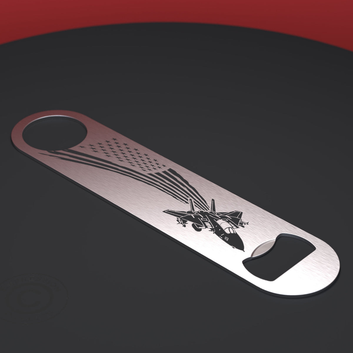 A stainless steel bottle opener with an engraved American flag and fighter jet design, displayed diagonally on a dark gray surface with a red background corner.