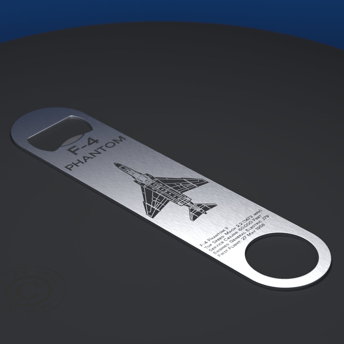 A silver metal bottle opener engraved with an F-4 Phantom jet fighter and the text 'F-4 PHANTOM', shown on a dark gray surface with a blue background in the upper corner.