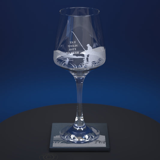 A clear glass wine glass with an etched  fishing scene and the words 'EAT SLEEP REPEAT' on the bowl, displayed on a square slate coaster against a dark blue background.