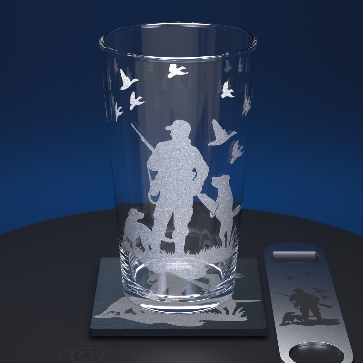 A clear glass pint tumbler with an etched hunting silhouette design, shown on a matching slate coaster next to a metal bottle opener with the same etched motif, against a dark blue gradient background.