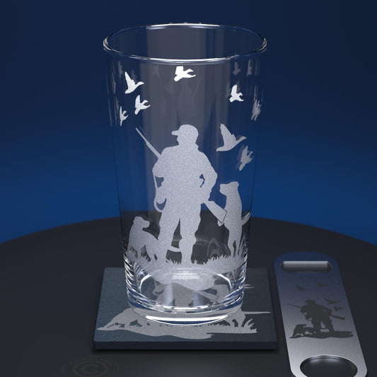 A clear glass pint tumbler with an etched hunting silhouette design, shown on a matching slate coaster next to a metal bottle opener with the same etched motif, against a dark blue gradient background.
