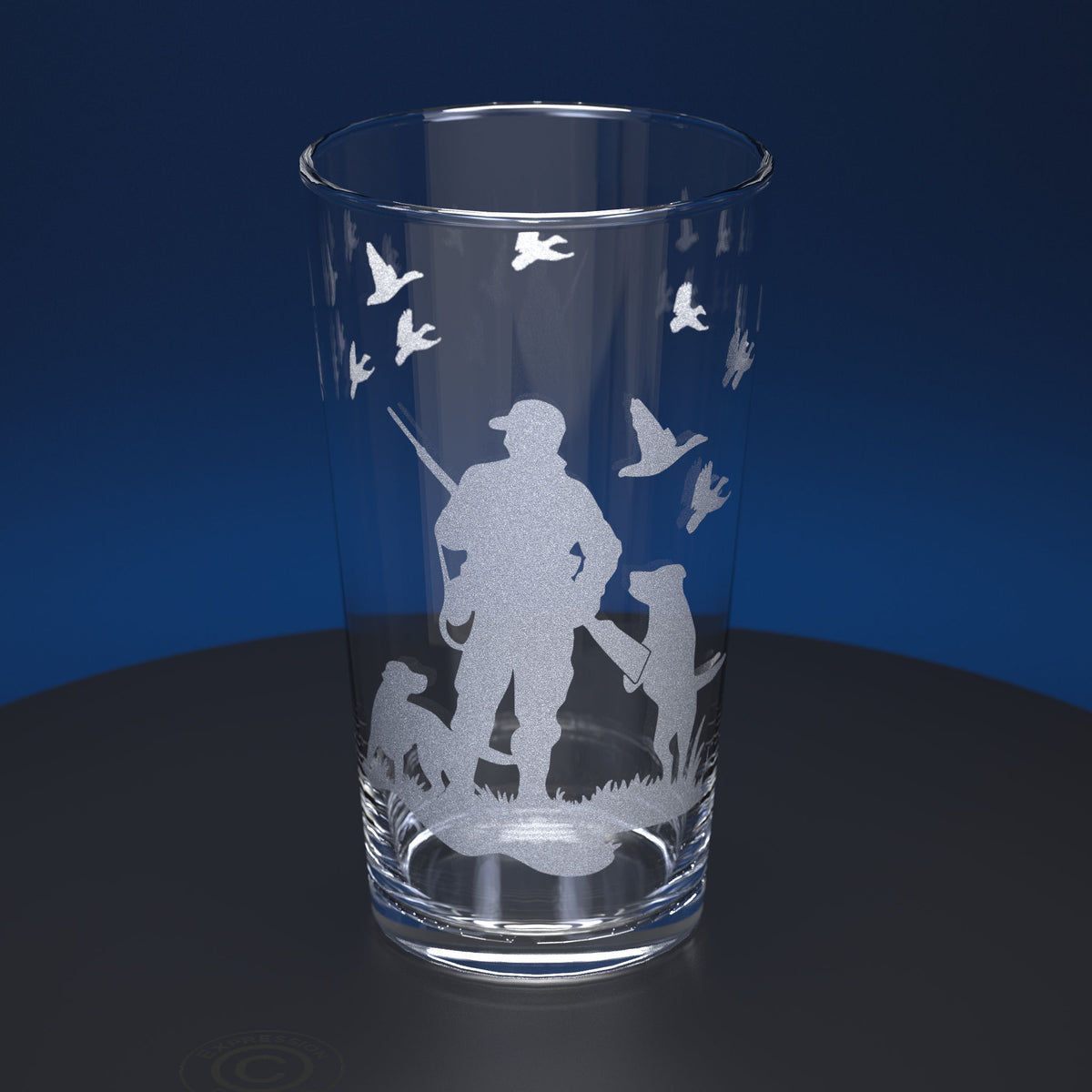 A clear pint glass with a frosted etched design of a hunting scene featuring a silhouetted hunter, two dogs, and several flying ducks, set against a dark blue background.