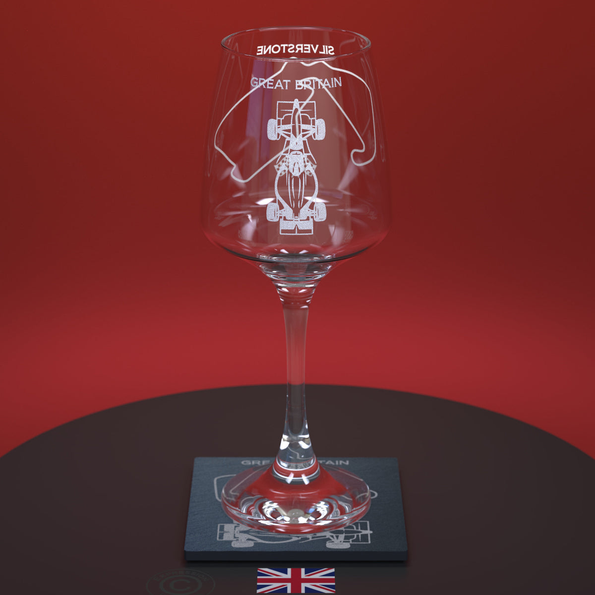 A clear stemmed wine glass with white etched graphics of a race car and the words 'SILVERSTONE' and 'GREAT BRITAIN', displayed on a dark coaster against a red background.